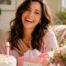 Happy woman celebrating her birthday at home with a cake, flowers, and birthday gifts in a bright sunlit room.