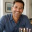 Smiling man celebrating his birthday at home, seated at a table with a birthday cake and wrapped gift in a bright, sunlit room.