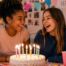 Two teenage friends laughing together at a birthday party with cake, candles, balloons, and gifts.