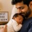 New father holding a sleeping newborn in a cozy living room on Father's Day, with a "Best Dad Ever" sign in the background.
